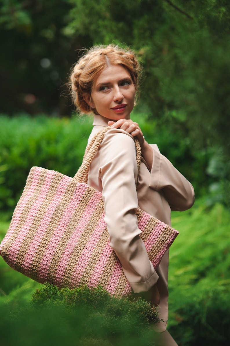 May include: A large, rectangular tote bag with alternating pink and tan stripes. The bag has a thick, woven texture and sturdy handles. The bag is carried over the shoulder, and the background is a lush green garden.