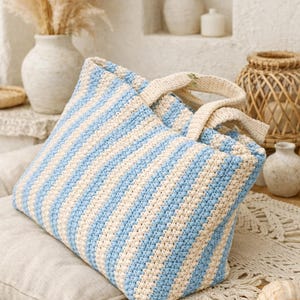 May include: A large, handmade crochet tote bag with alternating light blue and cream-colored stripes. The bag has sturdy cream-colored handles and is displayed on a neutral-toned cushion. The bag is a stylish accessory for carrying items.