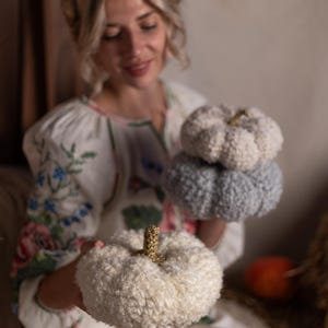 May include: A woman in a white dress with floral embroidery holds three crocheted pumpkins. The pumpkins are white, light blue, and a darker blue.