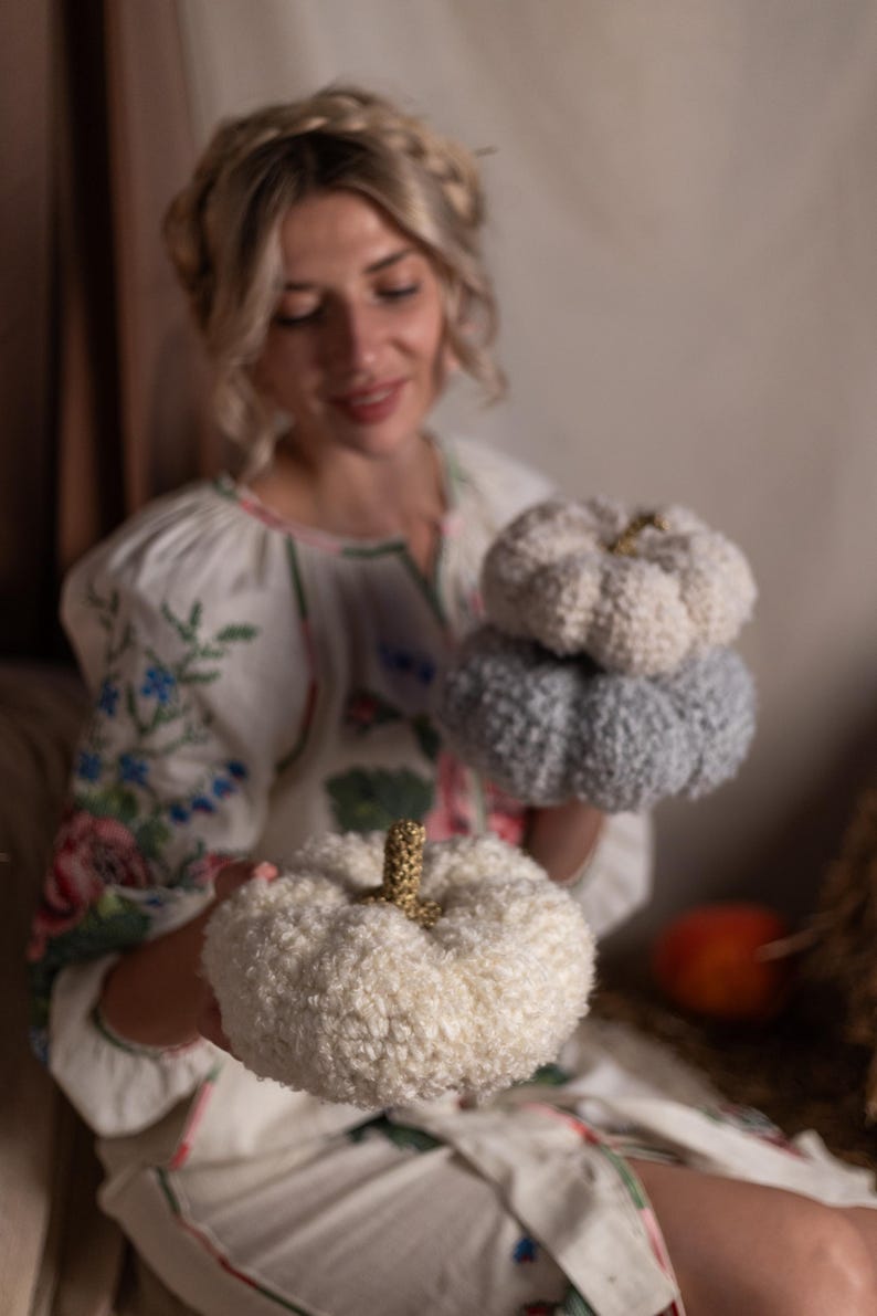 May include: A woman in a white dress with floral embroidery holds three crocheted pumpkins. The pumpkins are white, light blue, and a darker blue.