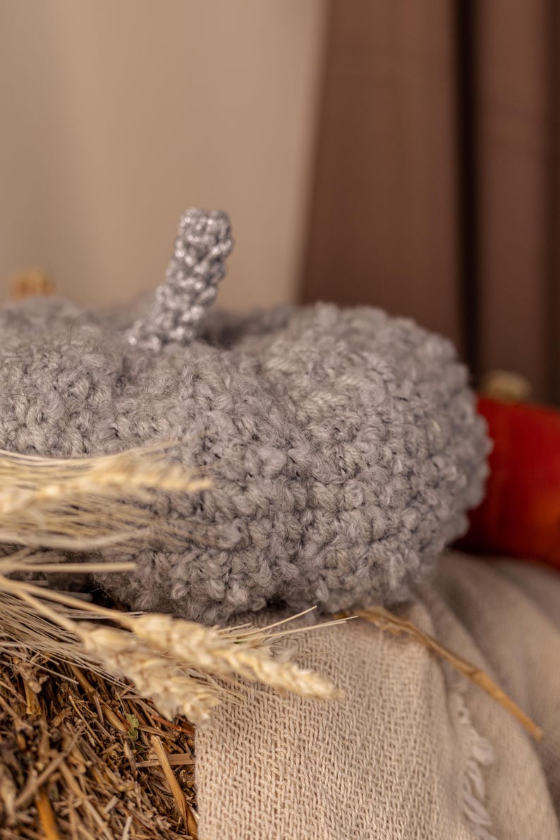 May include: A gray fuzzy pumpkin-shaped hat with a gray stem. The hat is sitting on a woven basket with dried wheat stalks.