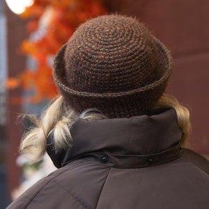 Può includere: Un cappello marrone all'uncinetto con tesa è indossato sopra una giacca marrone scuro. Il cappello è realizzato con un filato strutturato e ha un design circolare. I capelli biondi sono visibili sotto il cappello.