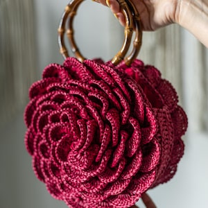 Puede incluir: Un bolso de mano de ganchillo burdeos con asa de bambú. El bolso tiene un diseño floral redondo. El asa está hecha de dos piezas de bambú curvadas. El bolso lo sostiene una persona.