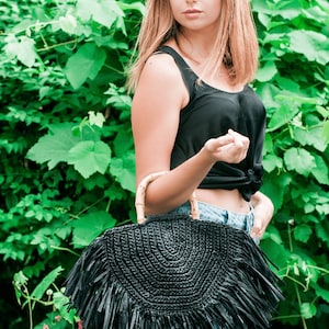 May include: A black, semi-circular woven handbag with a bamboo handle and fringe detail. The bag is held in front of a green leafy background. The bag's design is a stylish accessory.