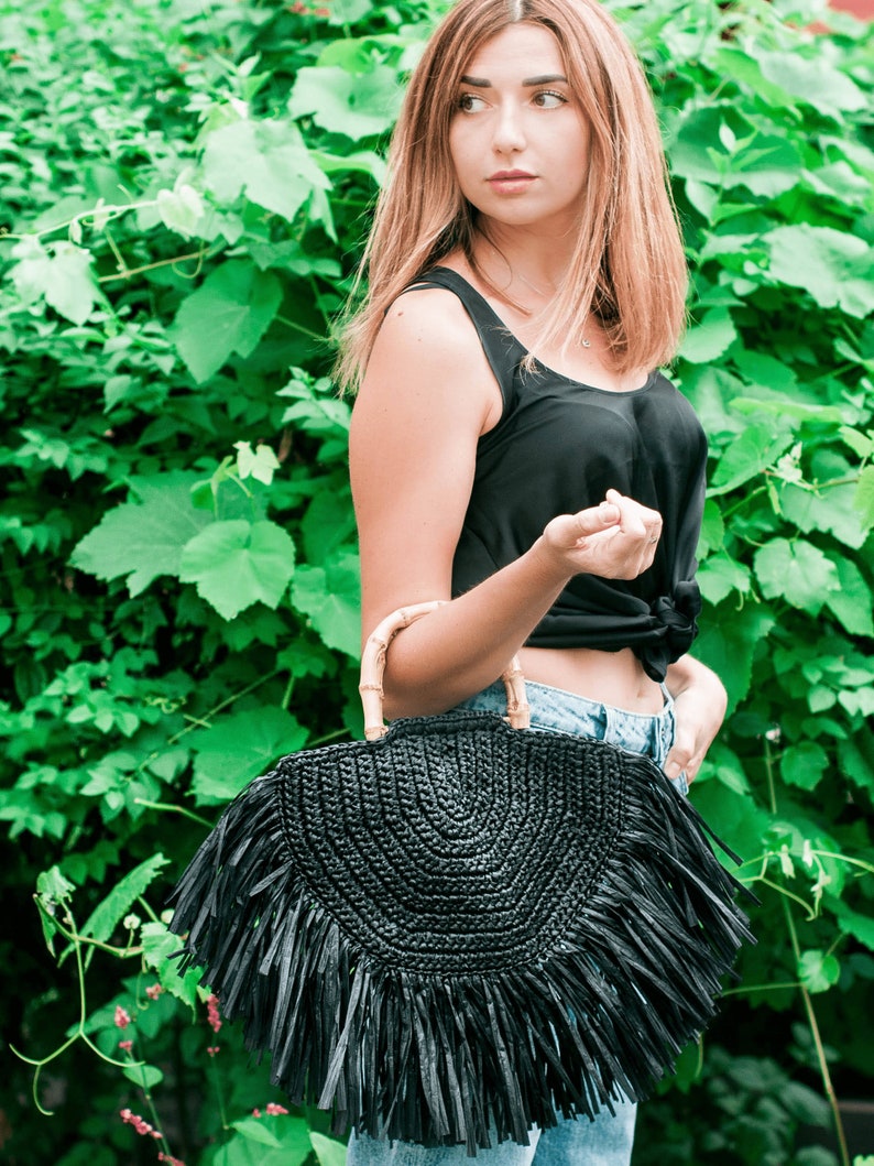 May include: A black, semi-circular woven handbag with a bamboo handle and fringe detail. The bag is held in front of a green leafy background. The bag's design is a stylish accessory.