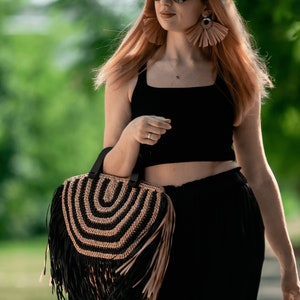May include: A black and tan fringed handbag with a circular design is held by a person. The bag has a black handle and a fringe that hangs down. The person is wearing a black top and pants, sunglasses, and large earrings.