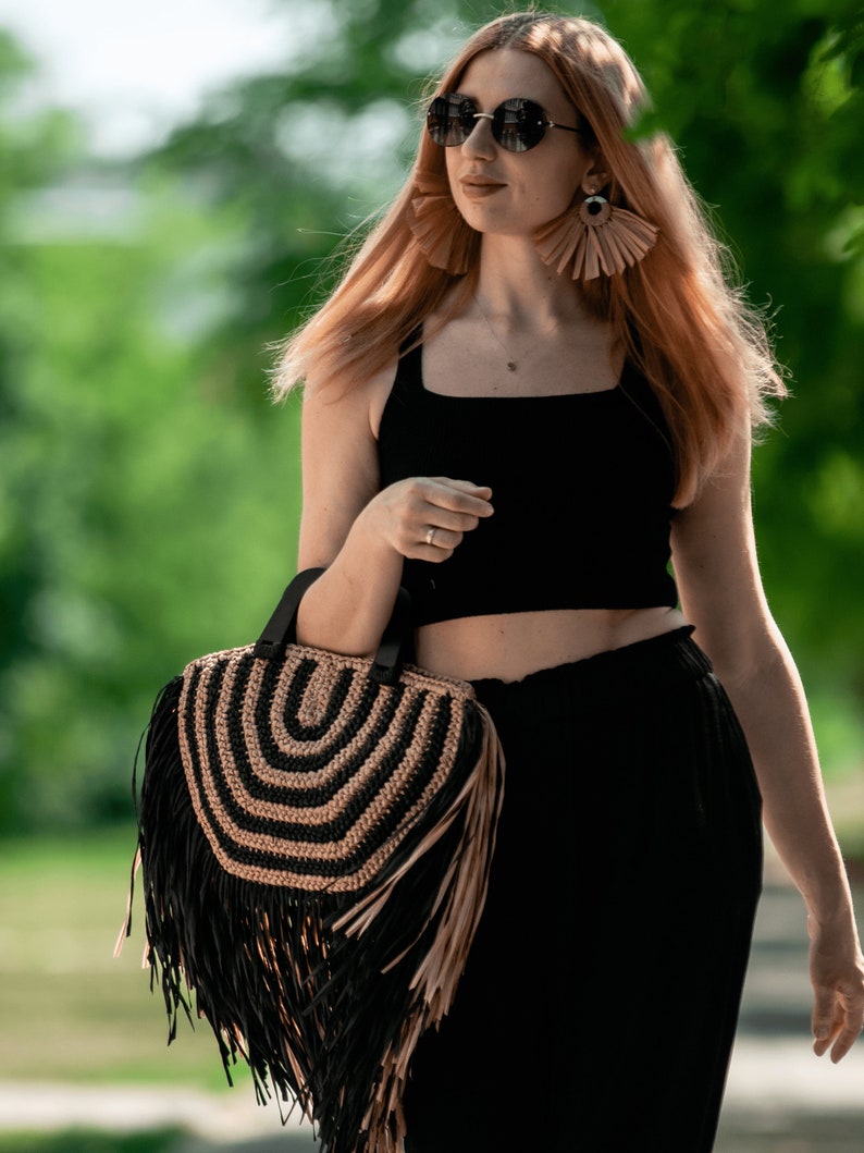 May include: A black and tan fringed handbag with a circular design is held by a person. The bag has a black handle and a fringe that hangs down. The person is wearing a black top and pants, sunglasses, and large earrings.