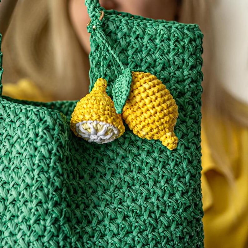 May include: A green woven tote bag with two yellow crochet lemon embellishments. One lemon is whole, the other is a half lemon. The bag has a woven texture and a matching green handle.