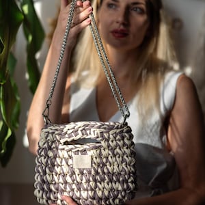 May include: A cream and lavender woven handbag with a square shape. It has a silver chain strap and a small label with text. The bag is held by a person. The bag's dimensions are in centimetres.