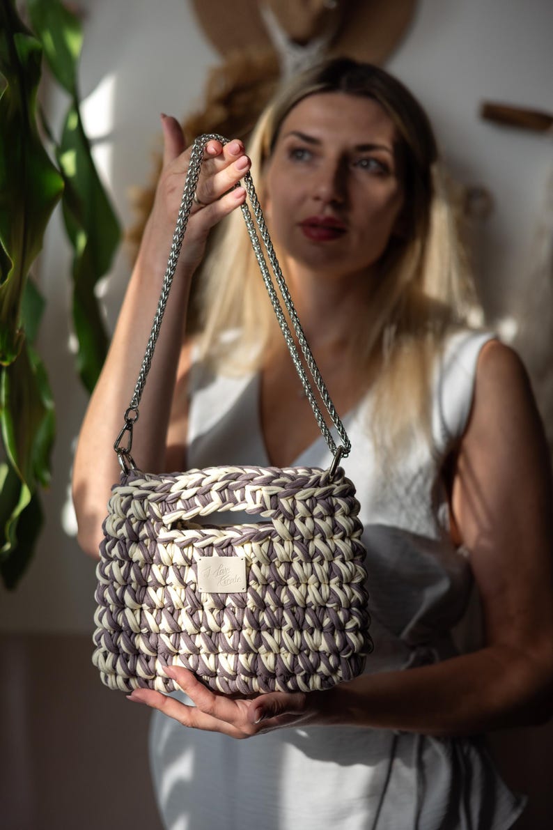 May include: A cream and lavender woven handbag with a square shape. It has a silver chain strap and a small label with text. The bag is held by a person. The bag's dimensions are in centimetres.