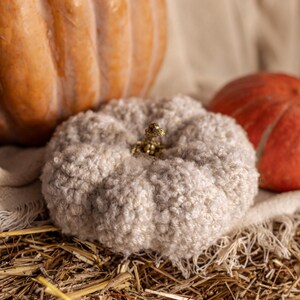 May include: A white crocheted pumpkin with a gold stem sits on a bed of straw with two other pumpkins in the background.