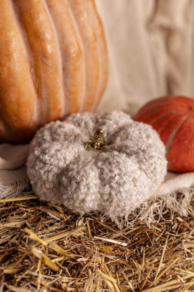 May include: A white crocheted pumpkin with a gold stem sits on a bed of straw with two other pumpkins in the background.