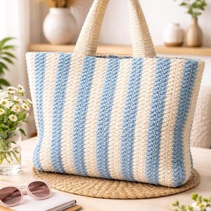 May include: A large, cream and light blue striped crochet tote bag with matching handles. The bag is rectangular and sits on a woven mat. Pink sunglasses and a notebook are on the table in front of the bag.