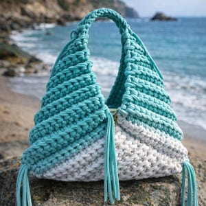 May include: A handmade, two-tone crochet bag in shades of turquoise and white, with a braided handle and turquoise tassels. The bag is sitting on a rock at the beach, with the ocean in the background.