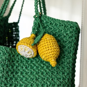 May include: A green woven tote bag with braided handles, decorated with two yellow lemon-shaped charms. One lemon is sliced, showing a white interior. The bag and charms are made of a textured material.