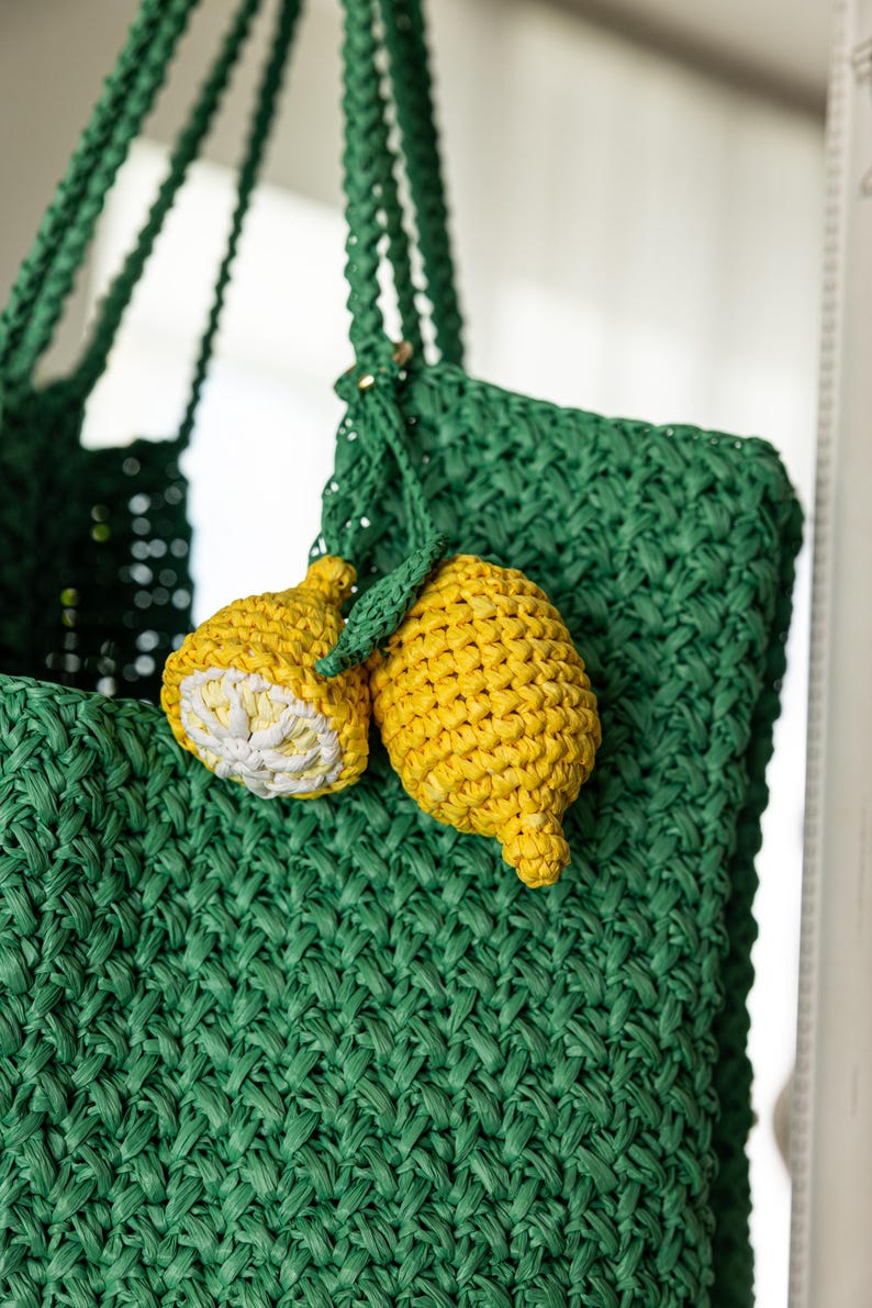 May include: A green woven tote bag with braided handles, adorned with two yellow lemon-shaped charms. One lemon is sliced, revealing a white interior. The bag and charms are made of a textured material.