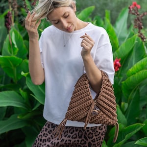 May include: A brown crochet handbag with a unique, textured design. The bag features a shoulder strap and decorative tassels. The woman in the image is wearing a white t-shirt and a leopard print skirt. The background is filled with green foliage.