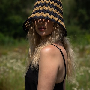 May include: A woman wearing a black tank top and a crocheted bucket hat with black and tan stripes. The hat is pulled down low over her forehead, and she is looking off to the side.