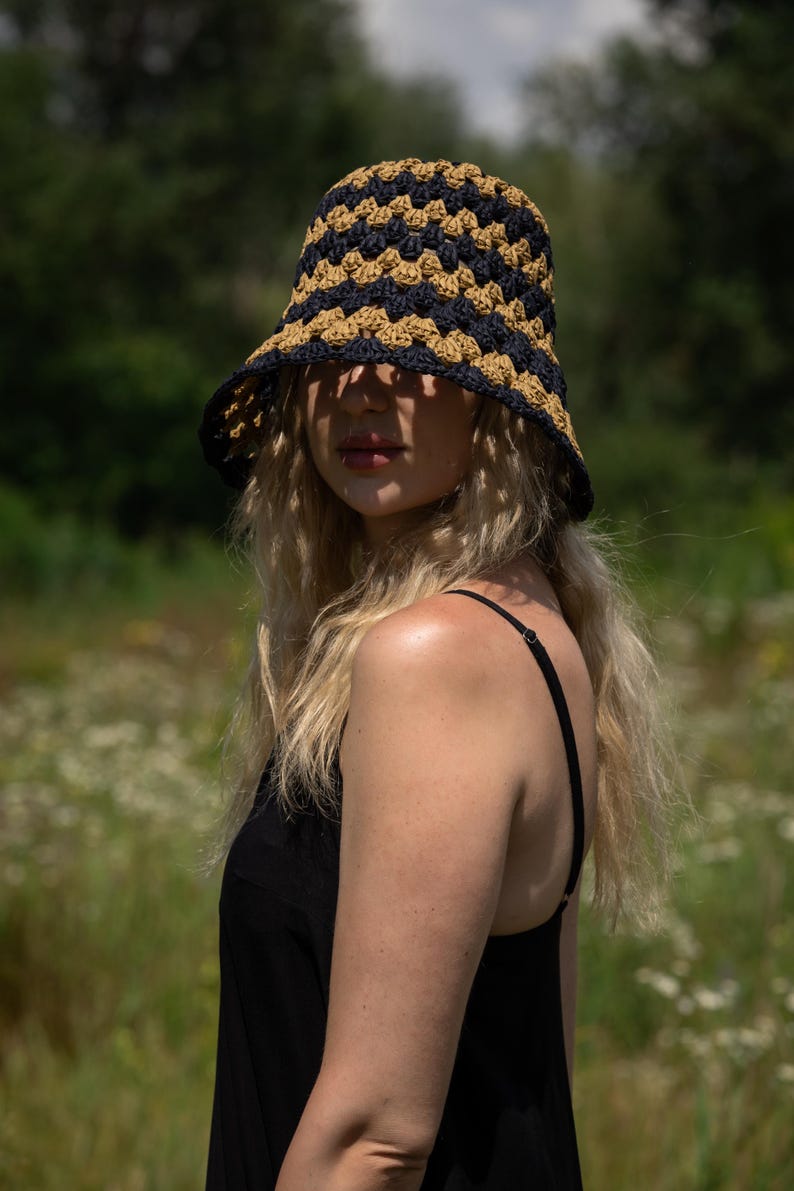 May include: A woman wearing a black tank top and a crocheted bucket hat with black and tan stripes. The hat is pulled down low over her forehead, and she is looking off to the side.