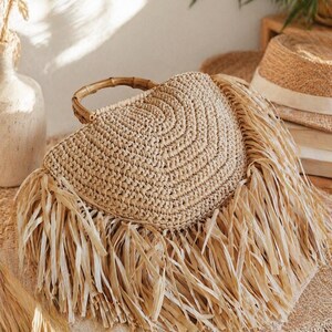 May include: A woven straw handbag with a circular design and bamboo handle. The bag features long, fringed edges and is in a natural beige color. The bag is a stylish accessory for summer.