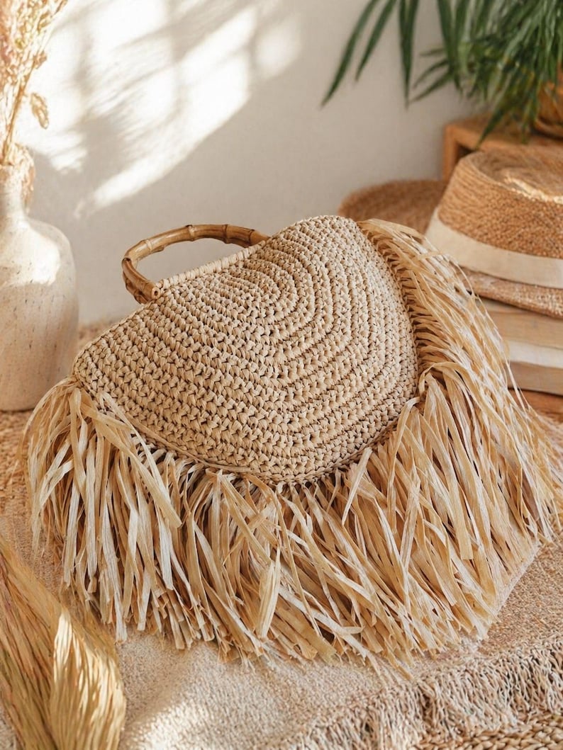 May include: A woven straw handbag with a circular design and bamboo handle. The bag features long, fringed edges and is in a natural beige color. The bag is a stylish accessory for summer.