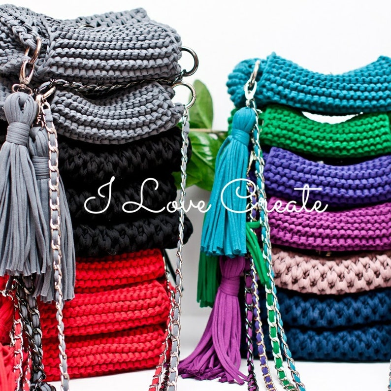 Puede incluir: Una pila de bolsos de crochet en varios colores, incluyendo gris, negro, rojo, azul verdoso, verde, morado, rosa y azul. Cada bolso tiene una correa de cadena plateada y un borla.