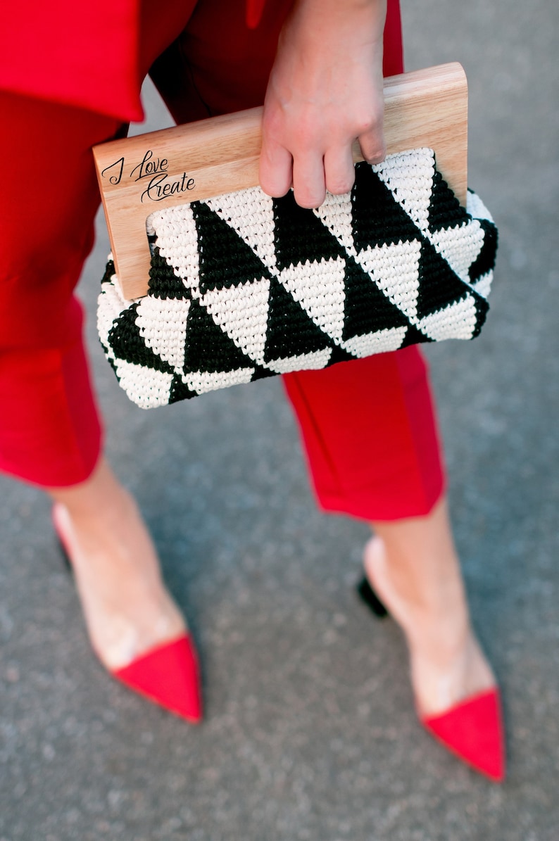 Op de afbeelding: Een clutch met een zwart-wit geometrisch patroon en een houten handvat. De tekst "I Love Create" staat op het houten handvat. De persoon draagt een rode broek en rode pumps met spitse neus.