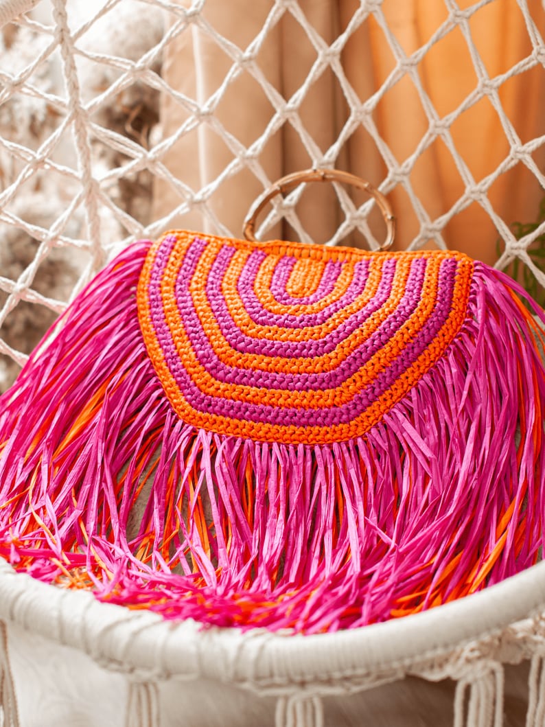 May include: A vibrant, handmade clutch with a half-circle design in shades of pink and orange. The clutch features a bamboo handle and is adorned with long, fringed edges. The bag is set against a white macrame backdrop.