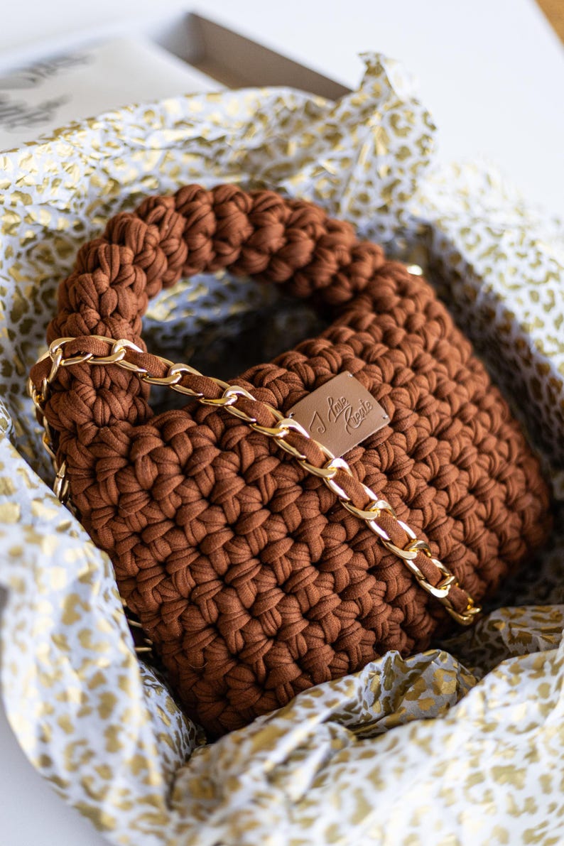 Op de afbeelding: Een bruine gehaakte handtas met een gouden kettingband. De tas heeft een klein metalen label met de tekst "I Love Croche"