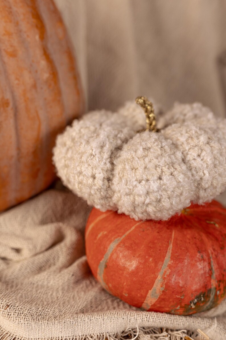 May include: A white fuzzy pumpkin sits on top of an orange pumpkin. Both pumpkins are on a beige fabric surface.