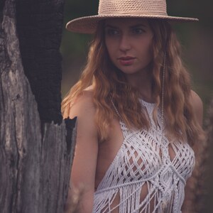 Peut inclure: Une femme portant un haut en crochet blanc avec une frange, un jean bleu et un chapeau de paille &agrave; larges bords avec un accent coquillage. Elle se tient devant un tronc d'arbre us&eacute;.