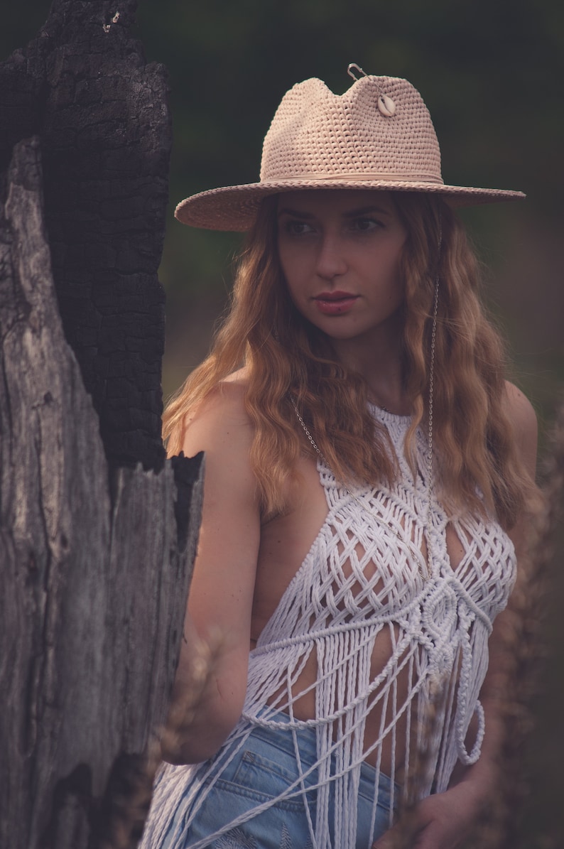 Peut inclure: Une femme portant un haut en crochet blanc avec une frange, un jean bleu et un chapeau de paille &agrave; larges bords avec un accent coquillage. Elle se tient devant un tronc d'arbre us&eacute;.
