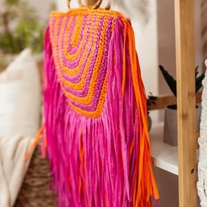 May include: A vibrant, handmade raffia bag with a bamboo handle. The bag features a woven orange and fuchsia design, with long fuchsia and orange fringe. The bag is a stylish accessory for carrying essentials.