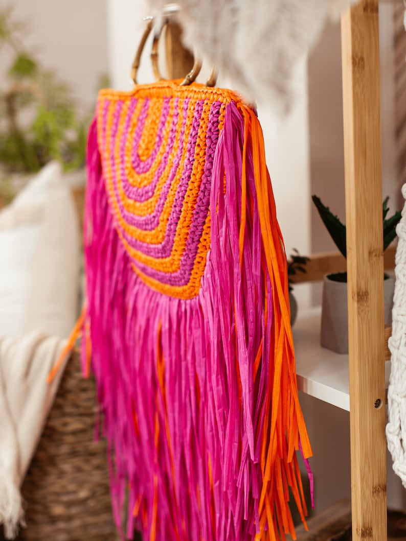 May include: A vibrant, handmade raffia bag with a bamboo handle. The bag features a woven orange and fuchsia design, with long fuchsia and orange fringe. The bag is a stylish accessory for carrying essentials.