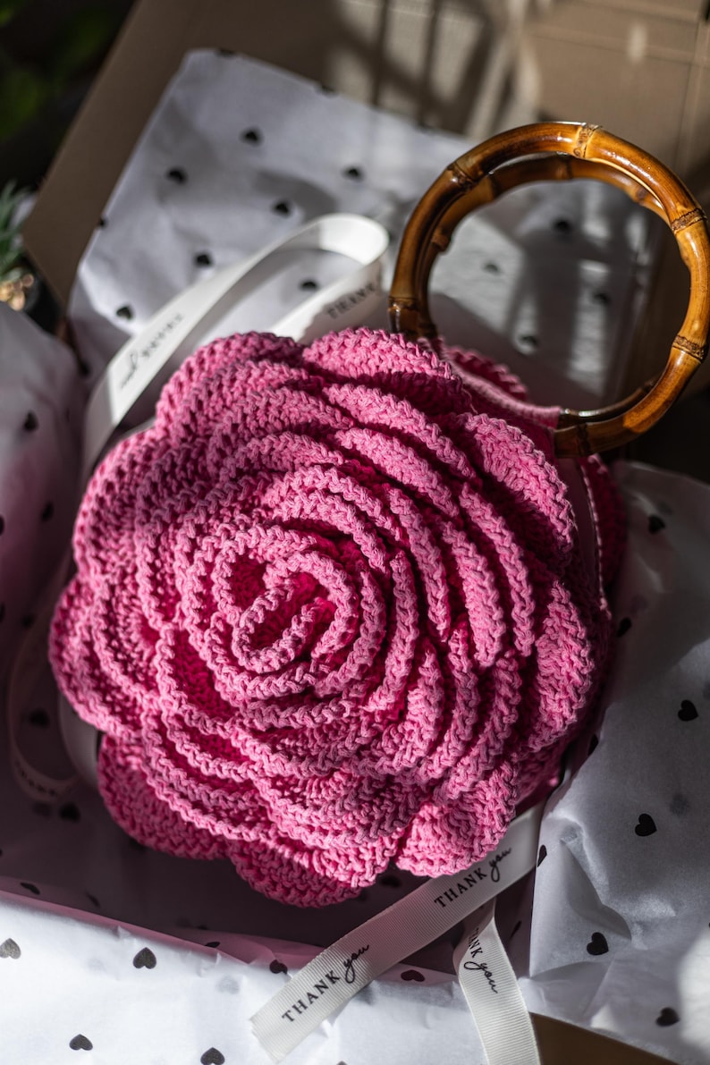 May include: A pink crocheted rose-shaped handbag with a bamboo handle. The bag is sitting in a box with white tissue paper and black hearts printed on it. The tissue paper has tags that say "Thank you".