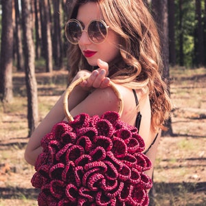 May include: A burgundy crocheted handbag with a bamboo handle. The bag has a unique, flower-like design. The woman in the image is wearing sunglasses and a black dress. The background is a forest with tall trees.
