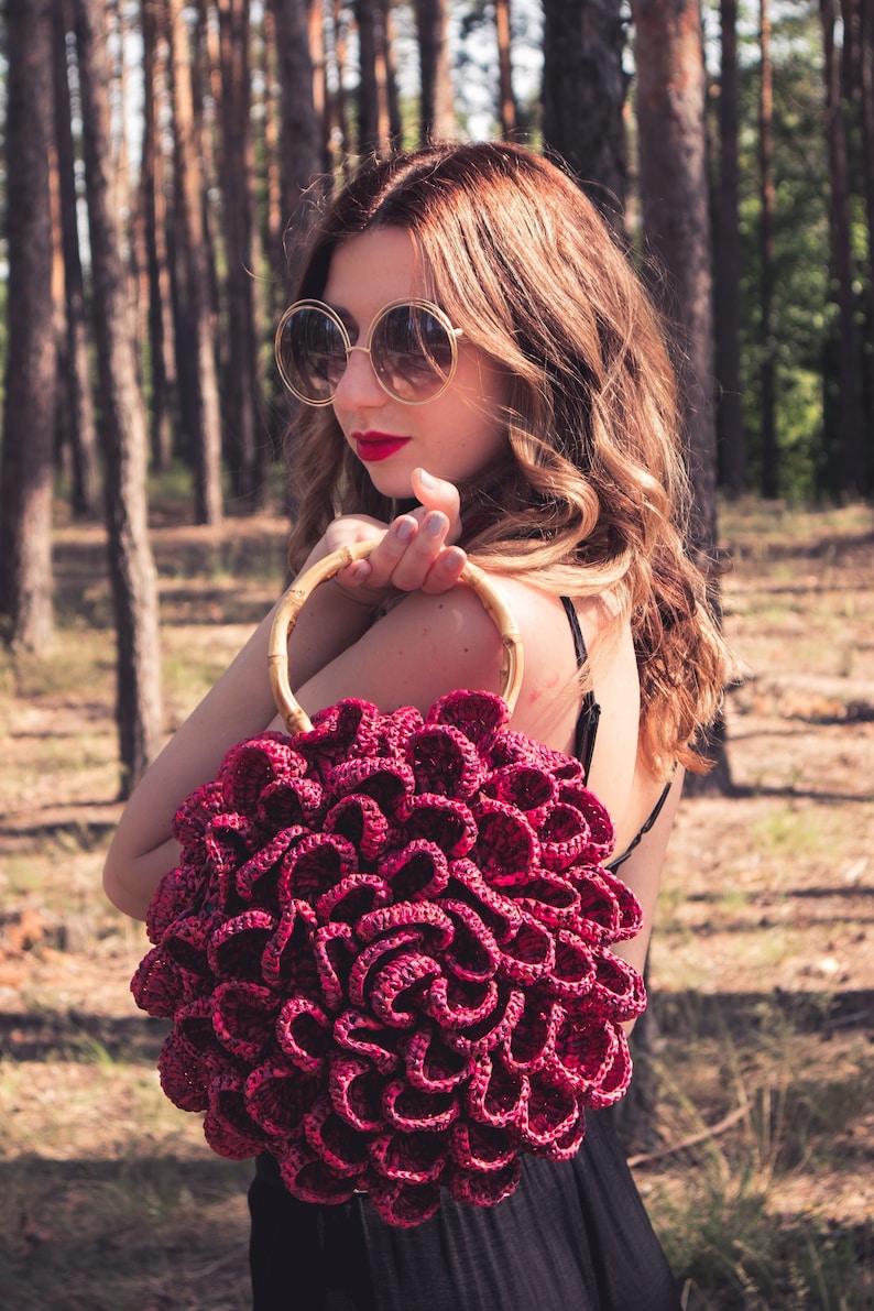 May include: A burgundy crocheted handbag with a bamboo handle. The bag has a unique, flower-like design. The woman in the image is wearing sunglasses and a black dress. The background is a forest with tall trees.
