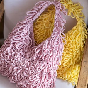 Könnte beinhalten: Zwei handgefertigte Taschen, eine rosa und eine gelbe, liegen auf einer weißen Oberfläche. Die Taschen sind aus geschlungenem Stoff gefertigt, wodurch ein strukturierter, gefranster Effekt entsteht. Die Taschen sind auf einem Holzstuhl.