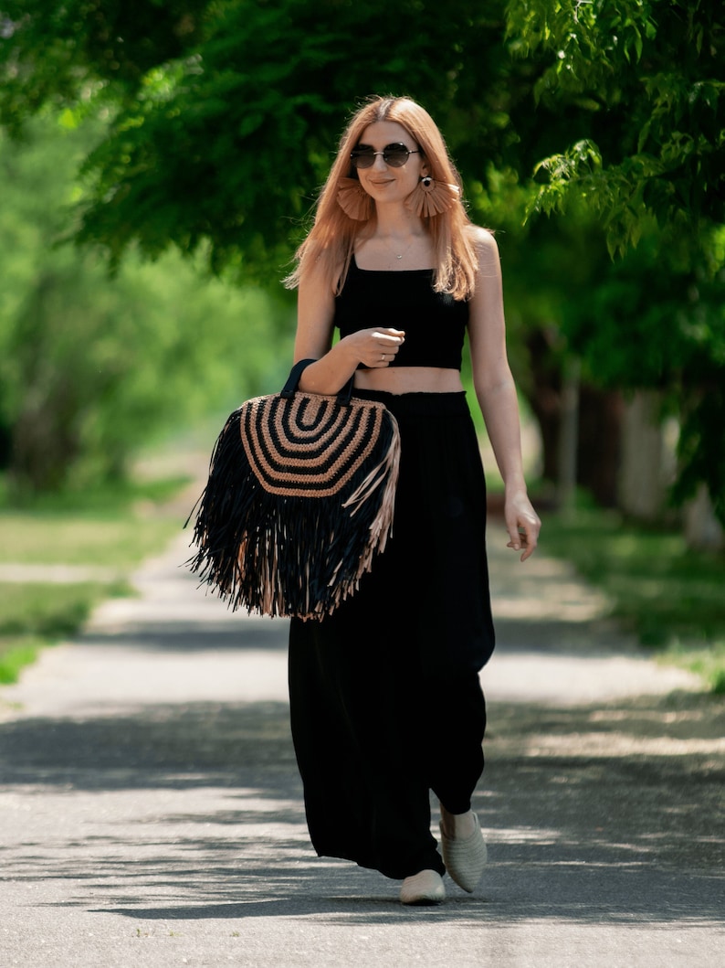 May include: A black crop top and wide-leg pants ensemble. A round straw handbag with black fringe and a woven design is carried. The person is wearing sunglasses and beige shoes, walking on a paved path.