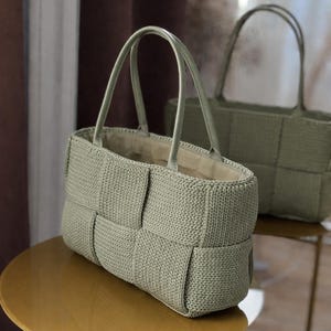 May include: Two sage green woven tote bags with leather handles sit on a gold table. The bags have a rectangular shape with a woven pattern. The handles are a matching sage green color. The background is blurred, suggesting an indoor setting.