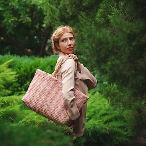 May include: A pink and tan striped tote bag with woven handles. The bag is carried over the shoulder against a backdrop of lush green foliage. The bag's design features alternating stripes of pink and tan.