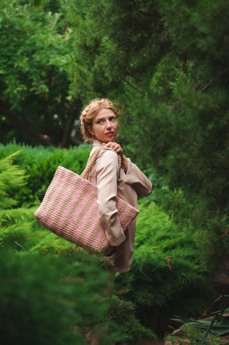 May include: A pink and tan striped tote bag with woven handles. The bag is carried over the shoulder against a backdrop of lush green foliage. The bag's design features alternating stripes of pink and tan.