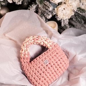 Op de afbeelding: Een roze gehaakte handtas met een witte gehaakte handvat. De tas heeft een klein label met de tekst "We Love Create".
