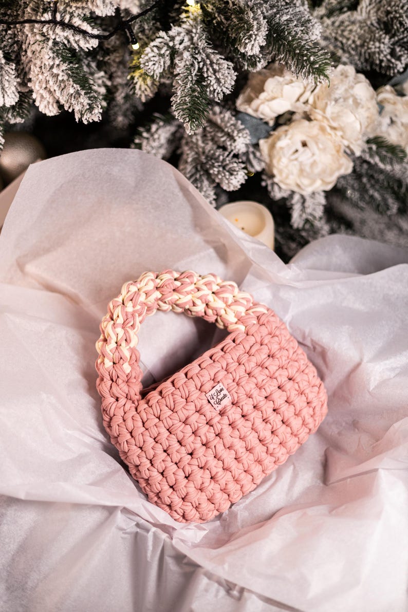 Op de afbeelding: Een roze gehaakte handtas met een witte gehaakte handvat. De tas heeft een klein label met de tekst "We Love Create".