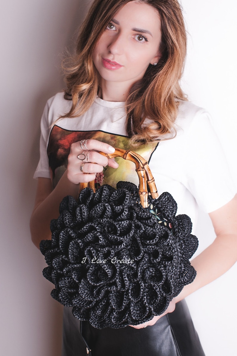 May include: Black crocheted handbag with a unique ruffled design. The bag features a bamboo handle and a small floral embellishment. The text "I Love Create" is visible on the bag.