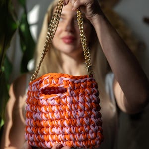 May include: A handmade crochet bag in orange and pink, with a gold chain strap. The bag has a rectangular shape with a large opening. The bag is held up by a person.