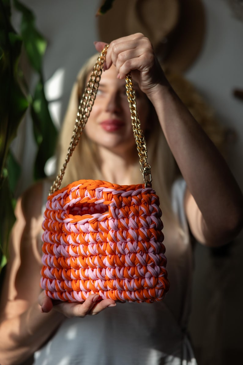 May include: A handmade crochet bag in orange and pink, with a gold chain strap. The bag has a rectangular shape with a large opening. The bag is held up by a person.