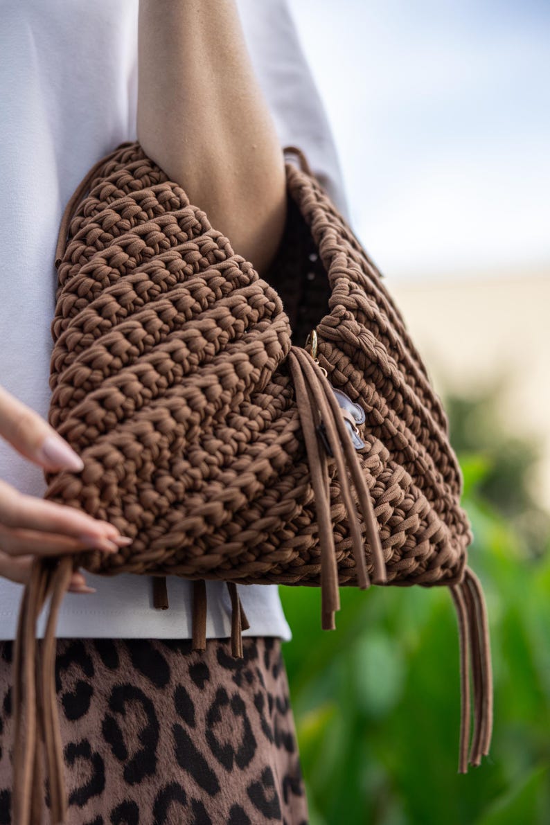 May include: A brown, hand-crocheted bag with a unique, textured design. The bag features a triangular shape and is adorned with long, dangling tassels. A small charm hangs from the side. The bag is held against a blurred background.