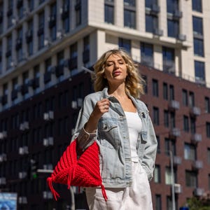 May include: A woman wearing a light blue denim jacket, white top, and white pants, carrying a bright red, hand-knitted bag. The bag has a unique, textured design and is the focal point of the image. The background features a modern building.