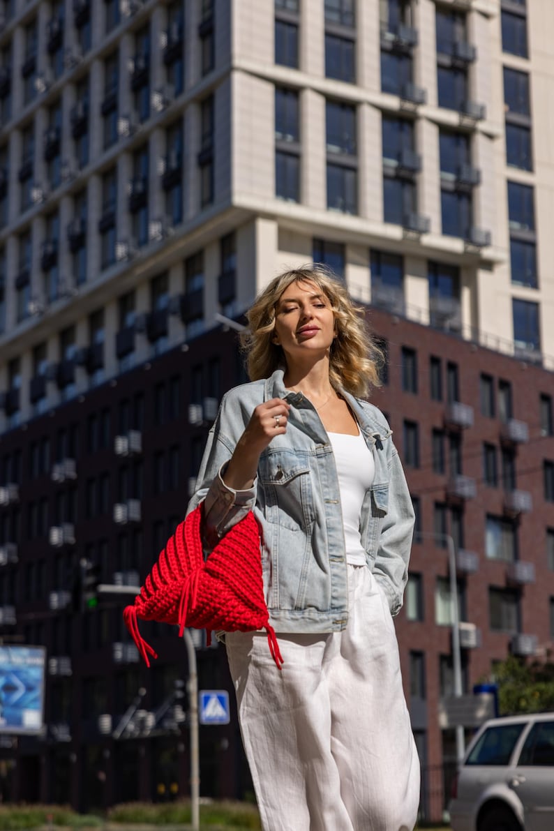 May include: A woman wearing a light blue denim jacket, white top, and white pants, carrying a bright red, hand-knitted bag. The bag has a unique, textured design and is the focal point of the image. The background features a modern building.